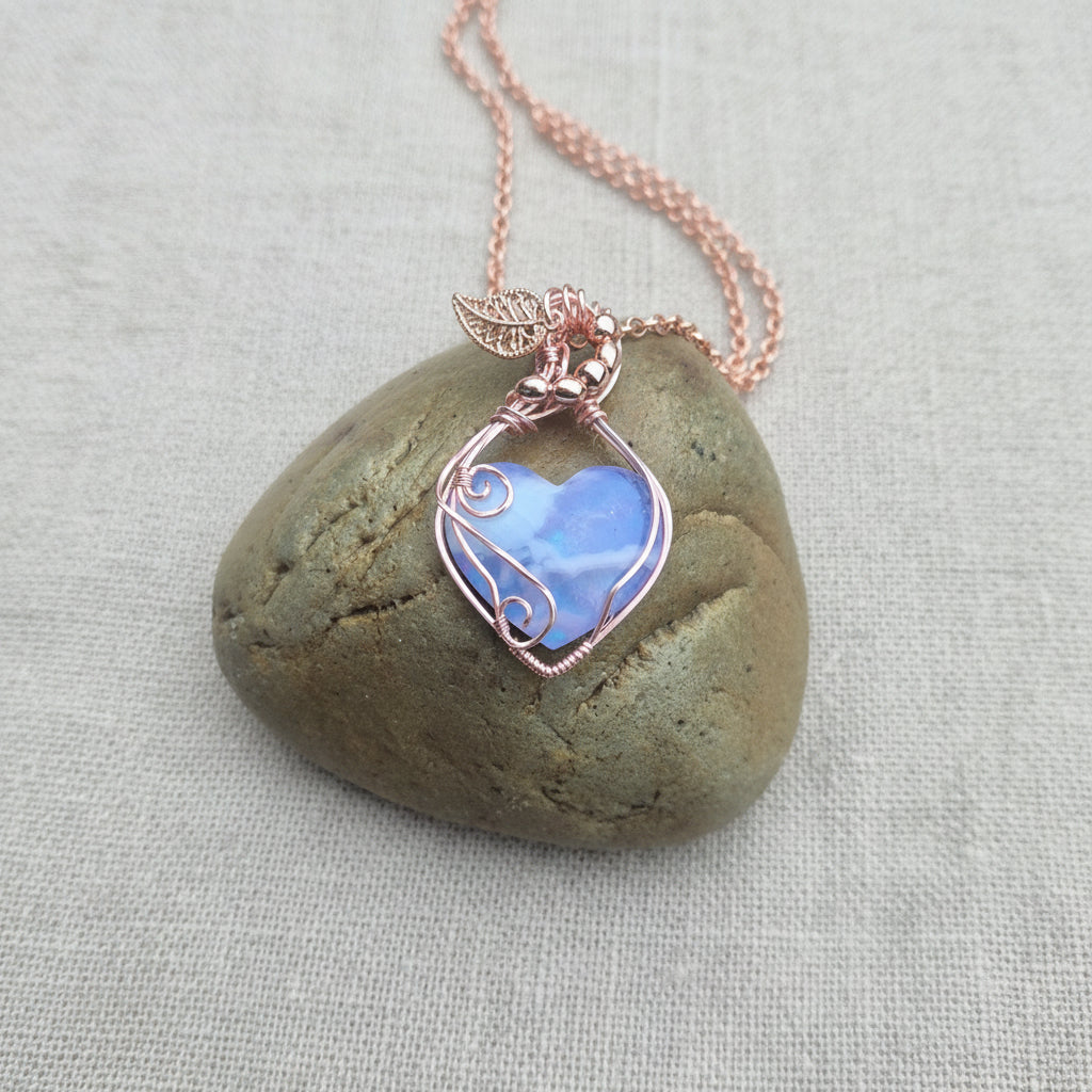 A heart shaped purple stone with cloud like patterns wrapped in whorls and swirls of rose gold coloured wire